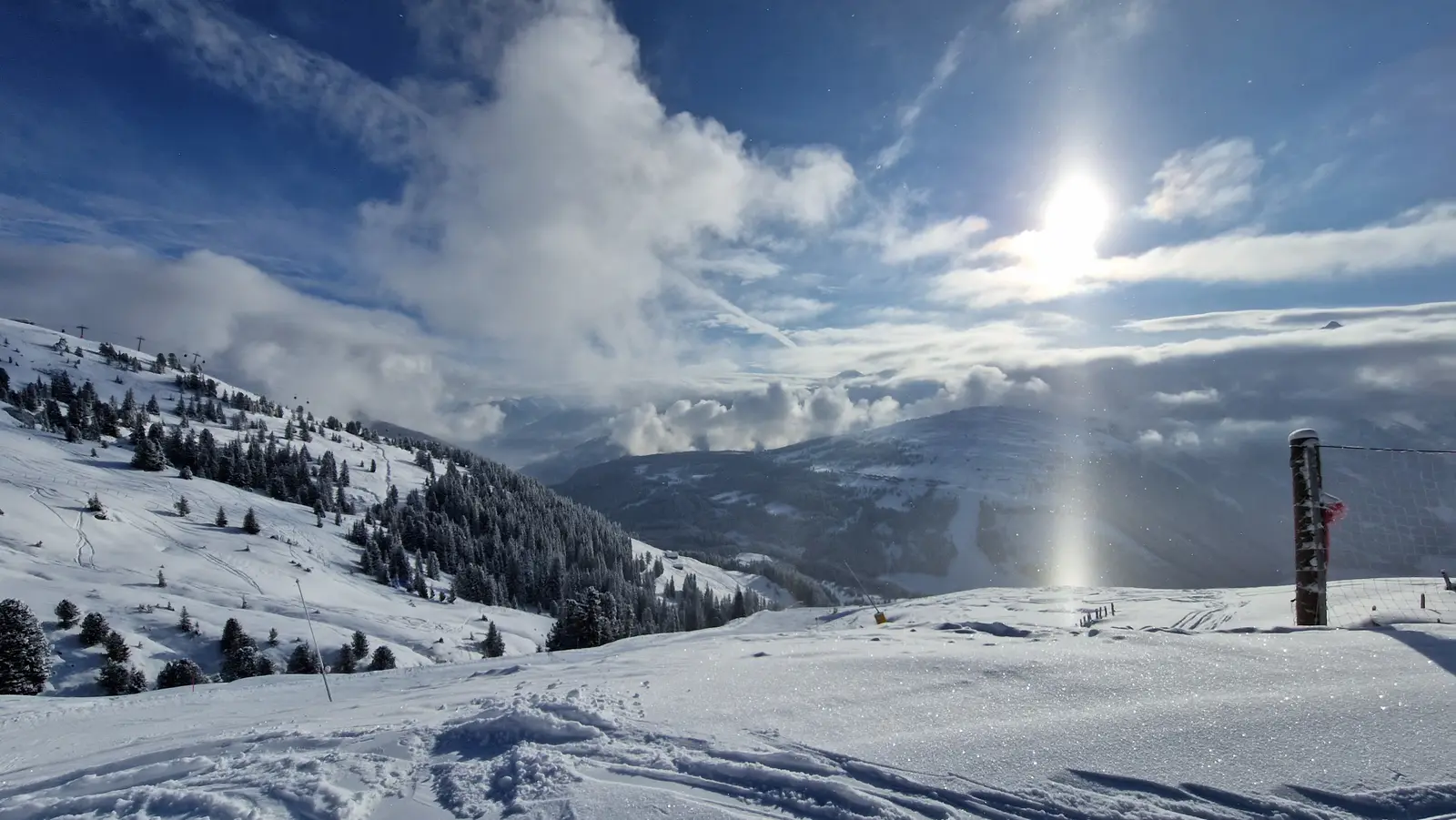 A sunlit, freshly snowed ski slope high above a sea of clouds, with snow-covered fir trees and mountains under a deep blue sky